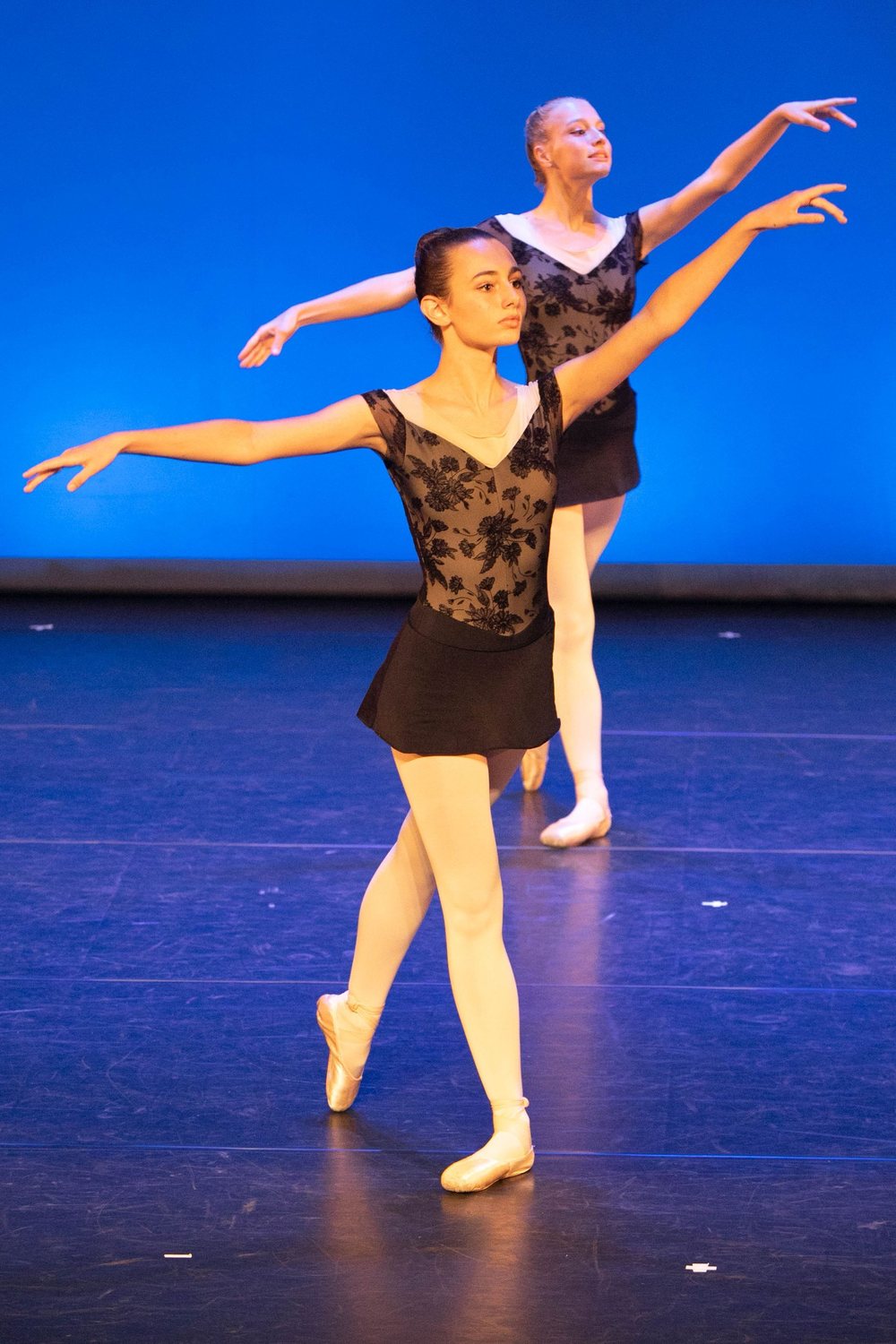 Deux danseuses sur scène, bras ouverts, en justaucorps noir, devant un fond bleu